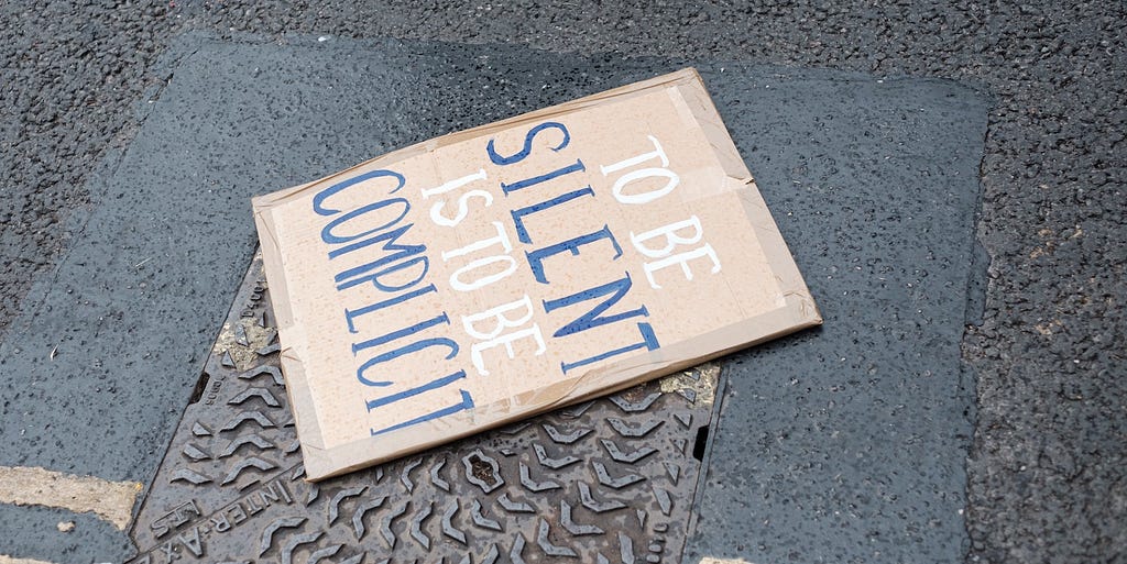 Image of a protest sign on the ground saying “to be silent is to be complicit”