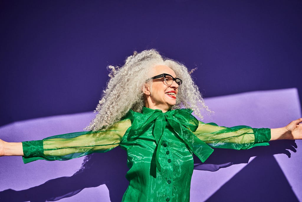 Portrait of a woman with gray curly hair and glasses wearing a green shirt against a purple background