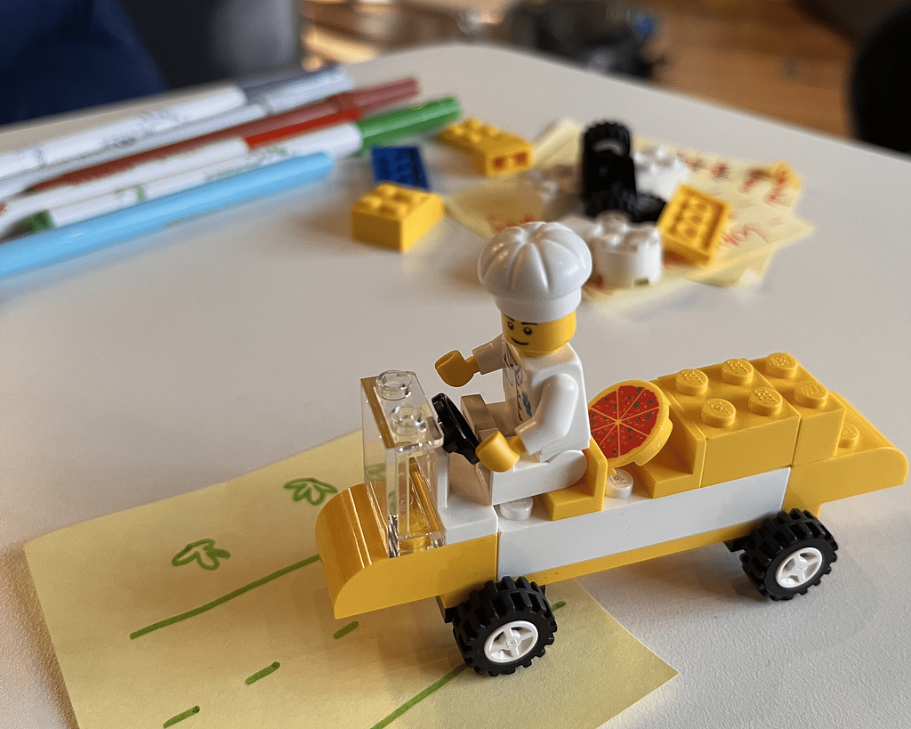 Imagem com o prototipo entrega para o cliente no nano desafio. Carro de lego nas cores amarelo e branco com um motorista entregador de pizza e uma pizza no branco traseiro.