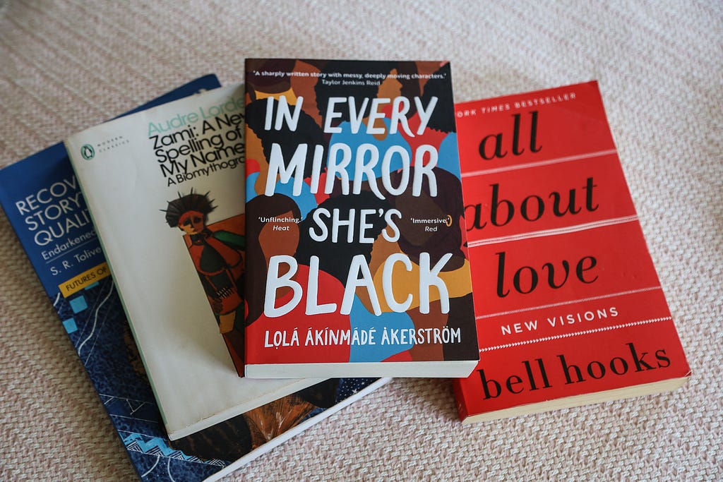 A selection of books on a fabric. The books are Recovering Black Storytelling in Qualitative Research: Endarkened Storywork by S.R. Toliver, Zami: A New Spelling of my Name by Audre Lorde, In Every Mirror She’s Black In Every Mirror She’s Black by Lola Akinmade Akerstrom and All About Love by Bell Hooks.