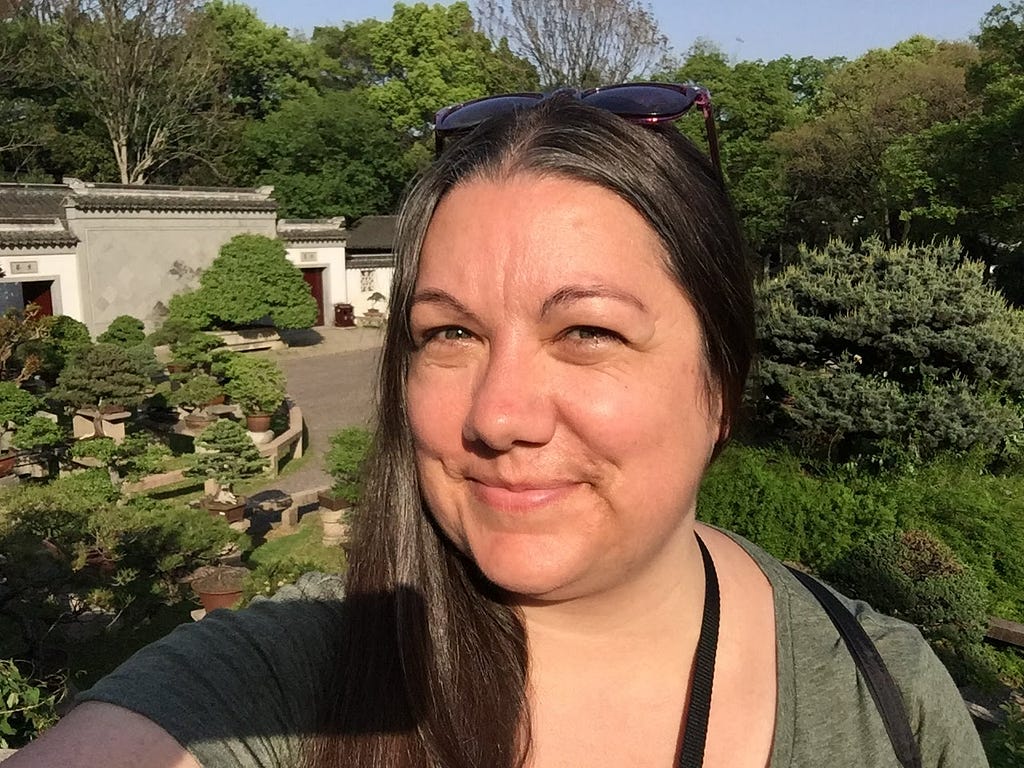 Woman with long brown hair pulled to one side, and sunglasses on her head, taking a selfie in Chinese garden.