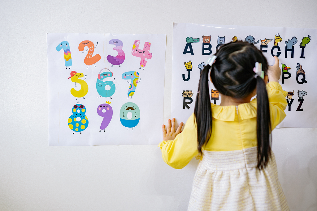 Image shows two charts, one containing numbers and the other containing letters. A preschool girl points at the chart containing letters, her back facing the viewer.