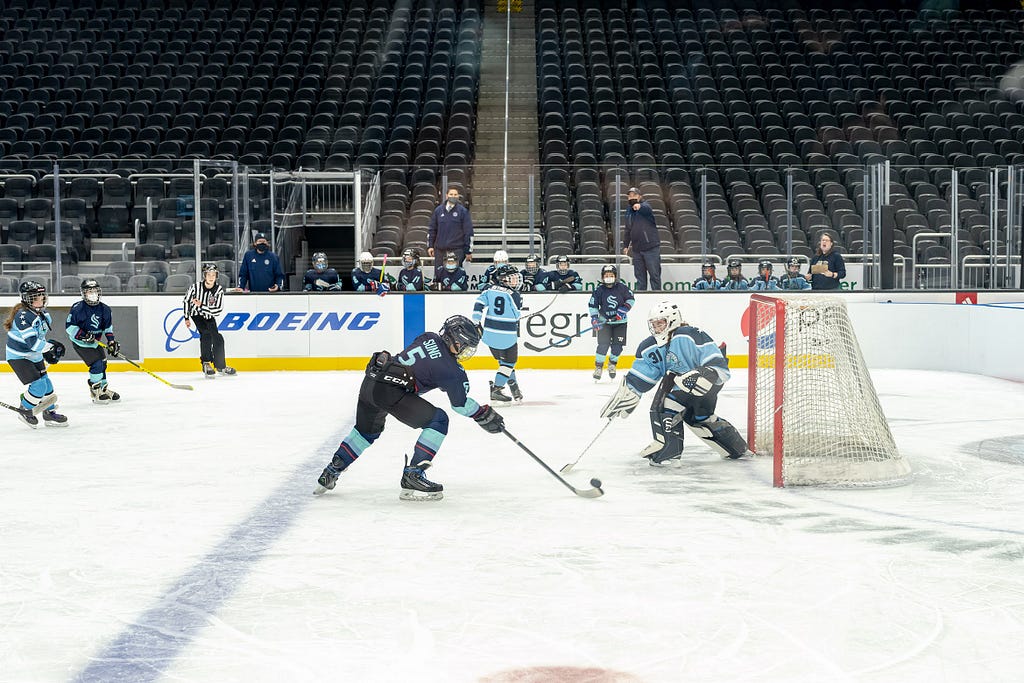 A hockey player shoots the puck while a goalie attempts to defend the goal. Teammates and a coach look on from behind the boards in the background.