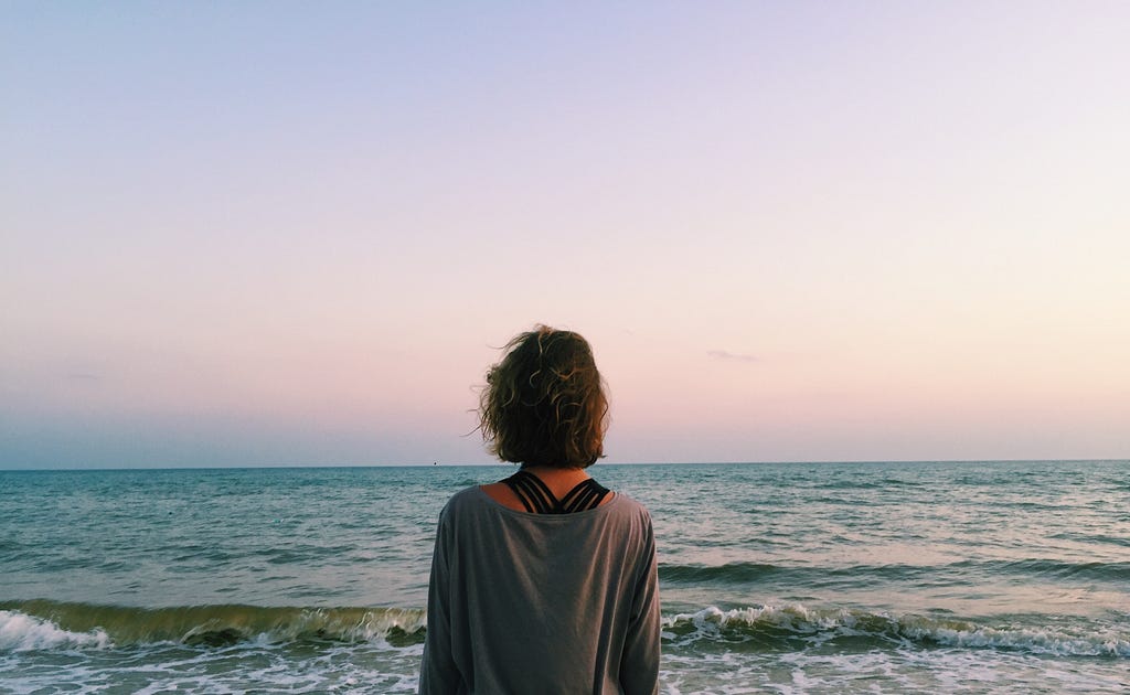 Money mindset: Woman standing with back to camera on beach, looking over the ocean and beautiful sun rise