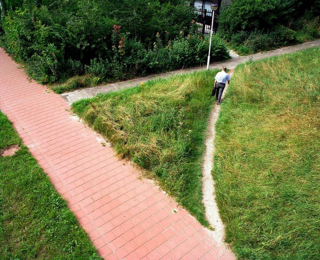 An aerial photo of a trodden path shortcutting two paved roads, called a ‘desire line’ by artist Jan Dirk van der Burg.