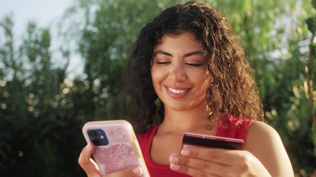 Young African American woman using cell phone holding credit card online shopping. Hispanic female shopper using instant easy mobile payments making purchase in online store. E-banking app service.