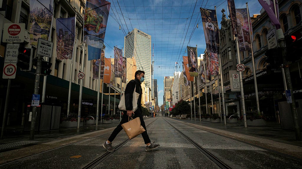 A photograph looking down a street in Melbourne. A person is crossing the road and tram tracks, carrying two bags and wearing a blue face mask. Along the street there are banners, street crossing signs, and traffic lights.