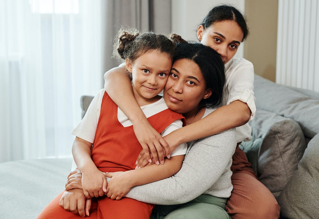 Two moms and a daughter sit on furniture and are shown embracing and looking into the camera.