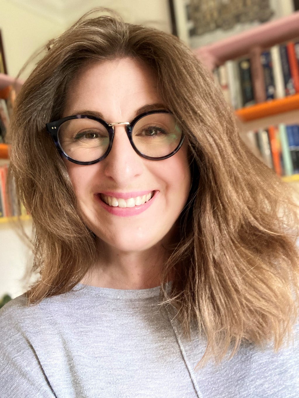 Photo of Viccy wearing large dark rimmed glasses and grinning, with orange and yellow bookshelves in the background