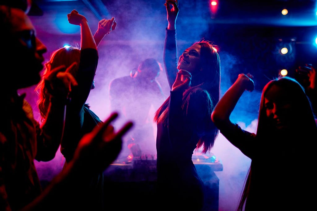 Silhueta de várias pessoas dançando com um ambiente escuro e o fundo iluminado com luzes azuis e roxas e uma fumaça.