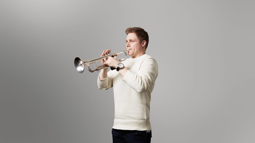 Philip Cobb playing trumpet wearing a white jumper, in front of a plain grey background