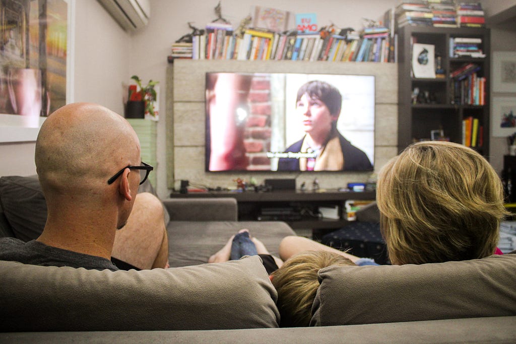 Família sentada no sofá, de costas para a foto, assistindo televisão na sala de estar de seu apartamento.