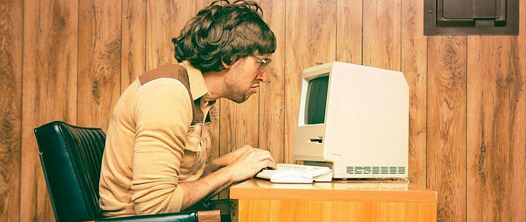 Image of a man with scruffy hair hunched over a vintage computer in a room with wood paneling. The man’s face is scrunched.