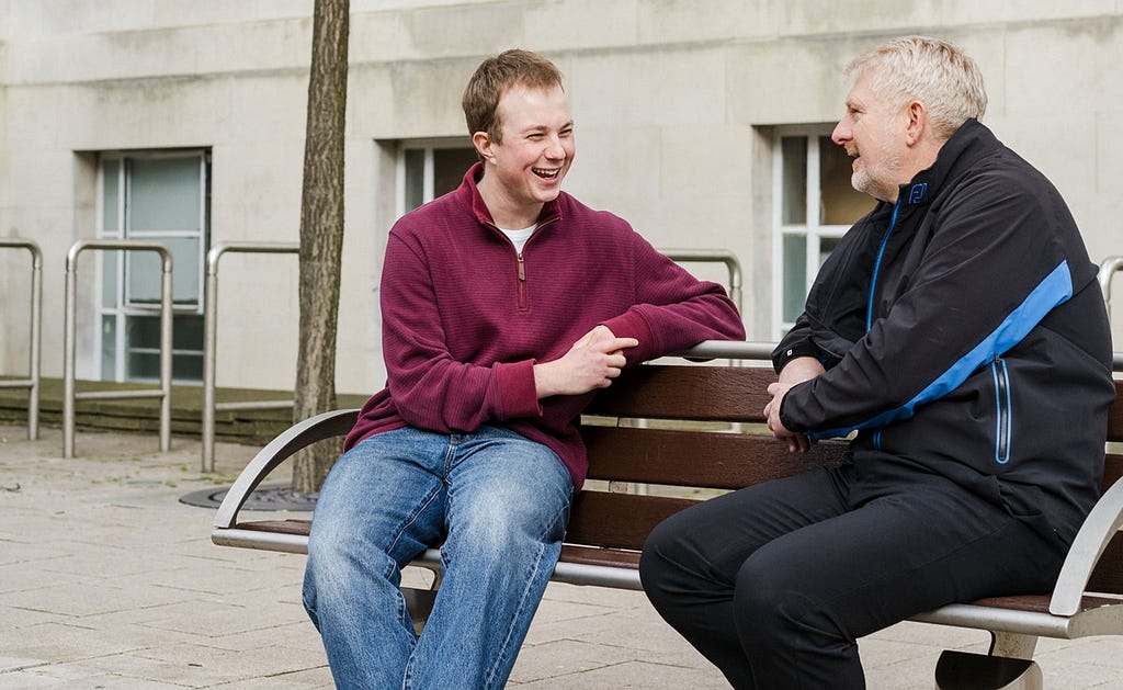 Jack is sitting on a bench outside with his mentor. They are both smiling while in conversation with each other.