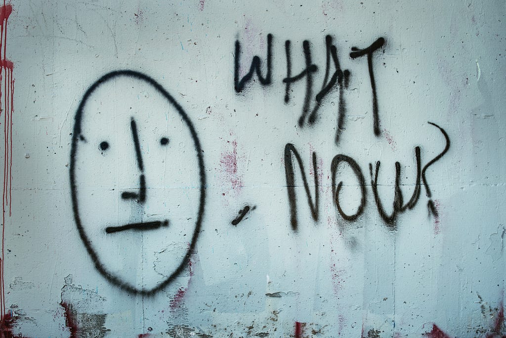 Graffiti on a white wall in black spray paint of a simple face with a neutral expression and text reading what now?