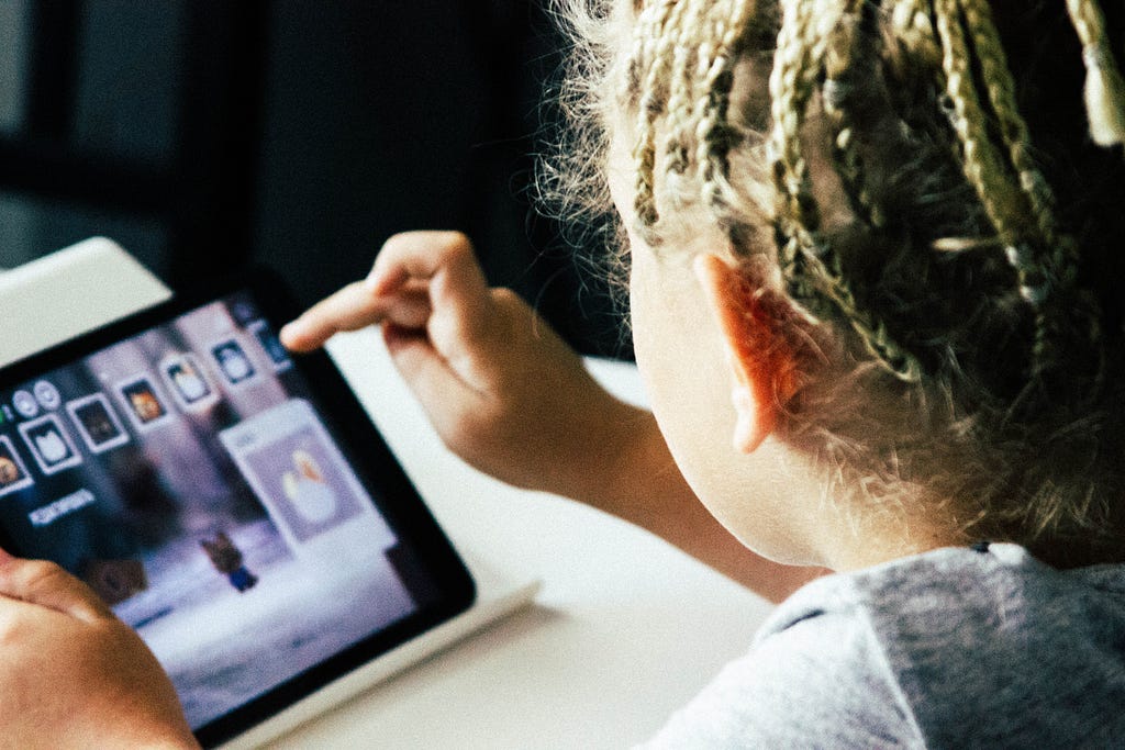 Meisje met veel dunne vlechtjes bediend het scherm van een tablet, waar ze met een spel bezig is.