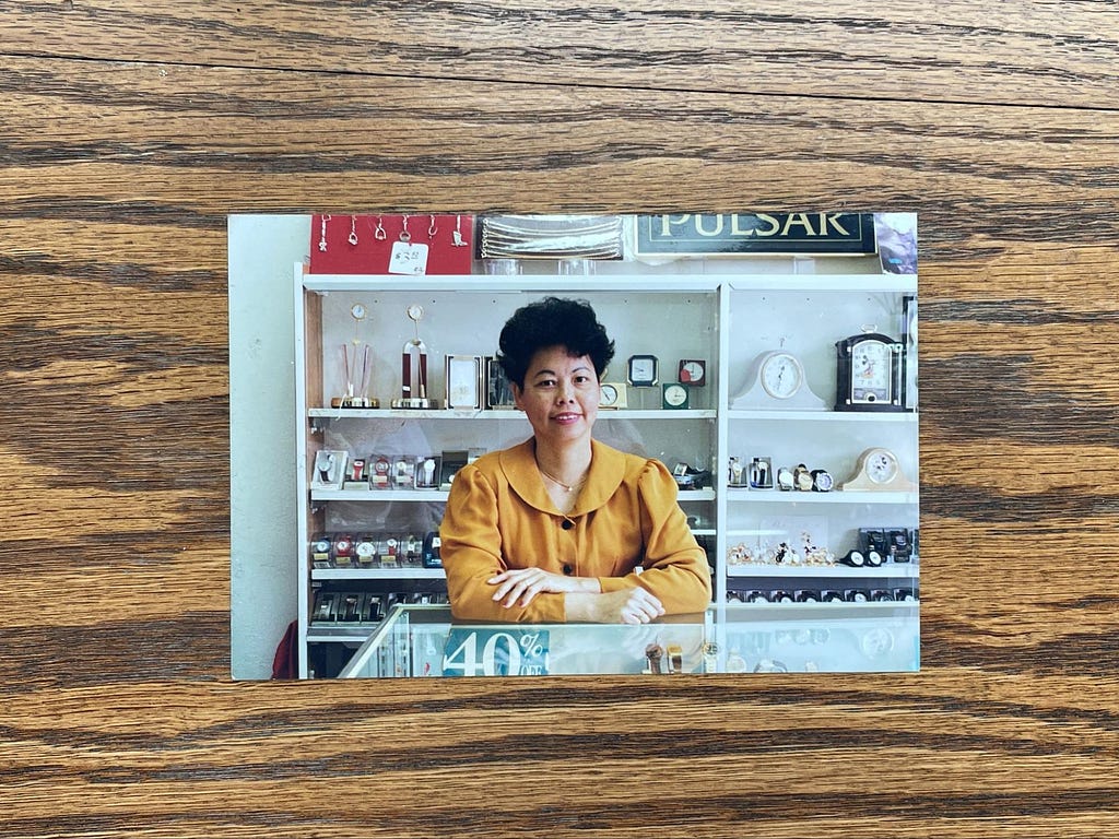 A photo of a printed photograph on a wood grain surface. The photo features Angela’s mom in a yellow blouse. She has blacked cropped hair and is wearing pink lipstick. She is leaning on the counter in her watch shop with her arms crossed and smiling at the camera. Behind her, there are shelves of various clocks, watches, and various merchandise.