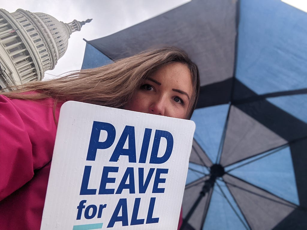 Woman with black and blue umbrella standing in front of US Capitol building holding a sign that reads “Paid Leave for All”