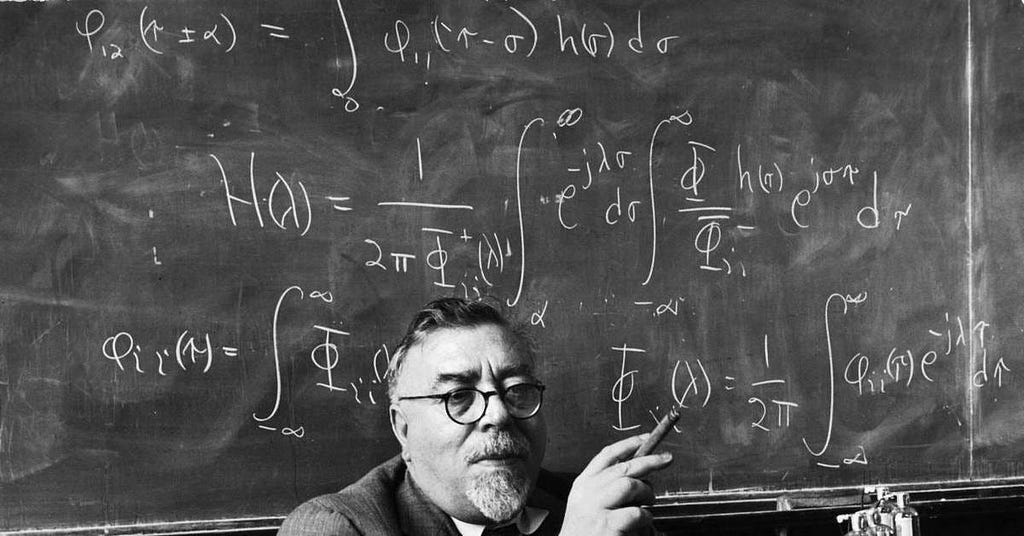 Black-and-white photo of Norbert Wiener seated in front of a chalkboard covered with complex mathematical formulas and integrals. He wears round glasses and a suit, holding a cigar in one hand while gesturing toward the equations with the other.