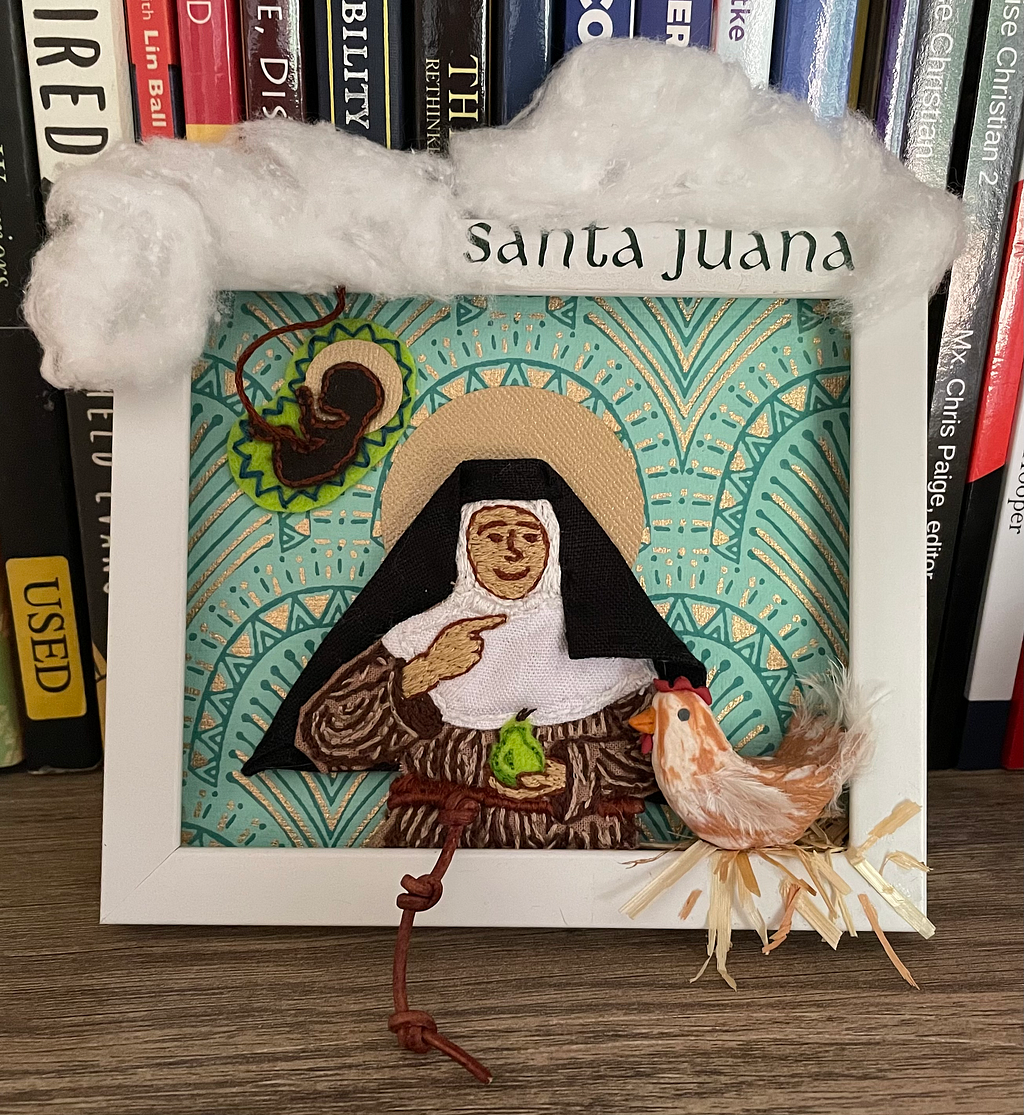 Multimedia artwork of Juana in a white rectangular frame. Juana wears her Franciscan robes, made with fabric and a leather belt; she smiles while pointing to her throat. Her other hand holds a green felt pear in front of her stomach. A polymer clay hen is perched on the frame in front of her, with straw glued beneath. To the upper left, a green felt womb holds a dark felt fetus with a gold halo, and an umbilical cord extending up into cotton clouds.