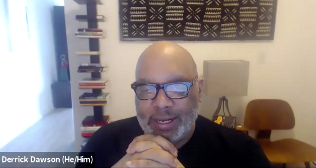 Zoom screenshot. Derrick. Black man. Black rimmed glasses, black shirt, bald, gray beard. His hands are clasped mid-sentence.