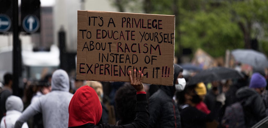 Protestor holding a cardboard sign that reads “It’s a privilege to educate yourself about racism instead of experiencing it!”