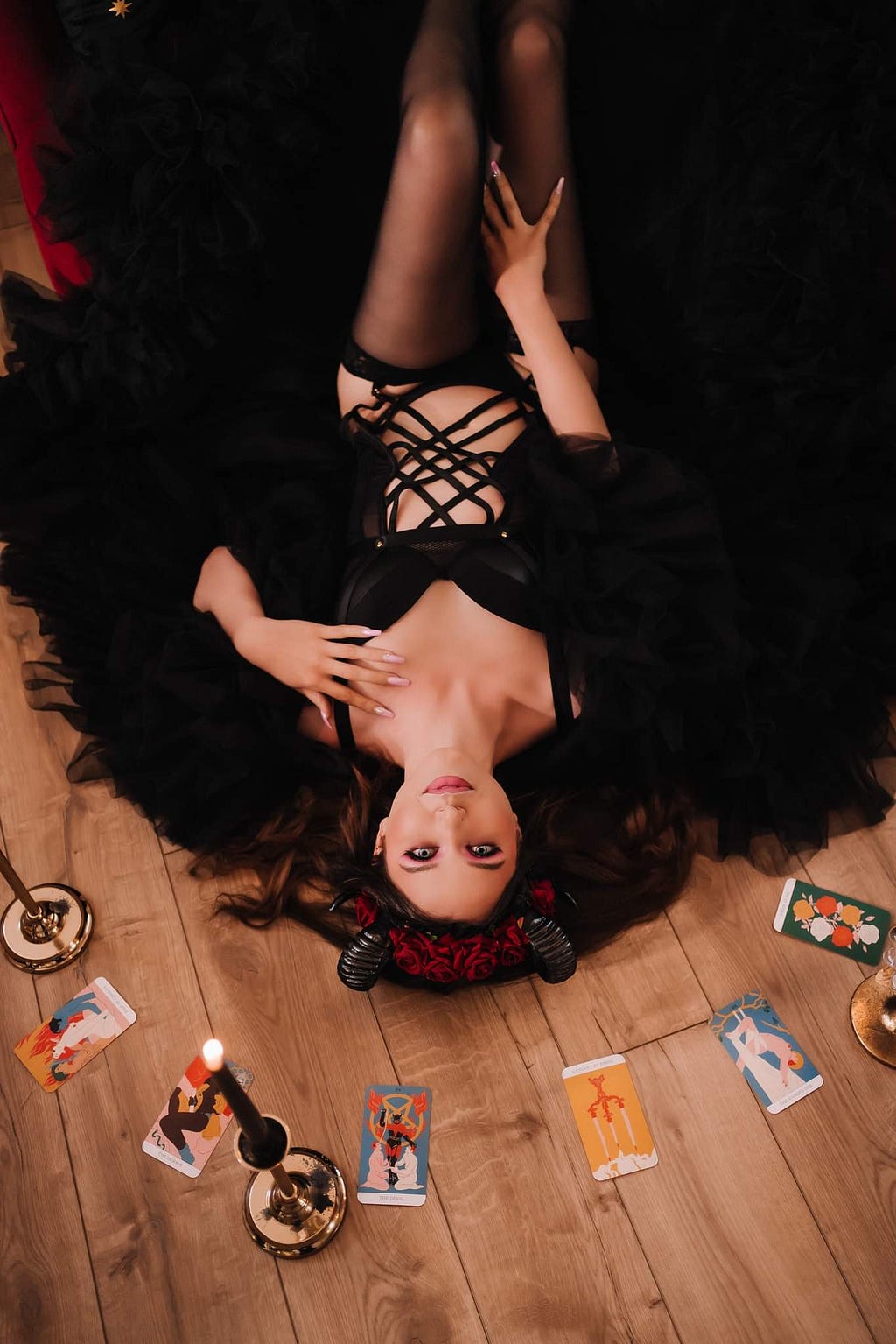 A thin white woman in black lingerie, wearing curved black horns, lays on the floor with tarot cards fanned out around her.