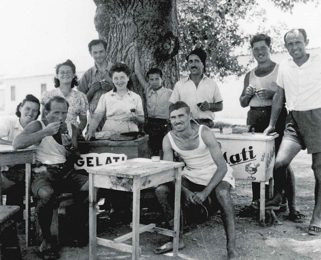 Seven men and three women group together in front of a wooden table and two ice cream carts. A man in the center of the image is smiling and wearing a white tank top and shorts. To his left, another man in a white tank top and shorts takes a bite of ice cream.