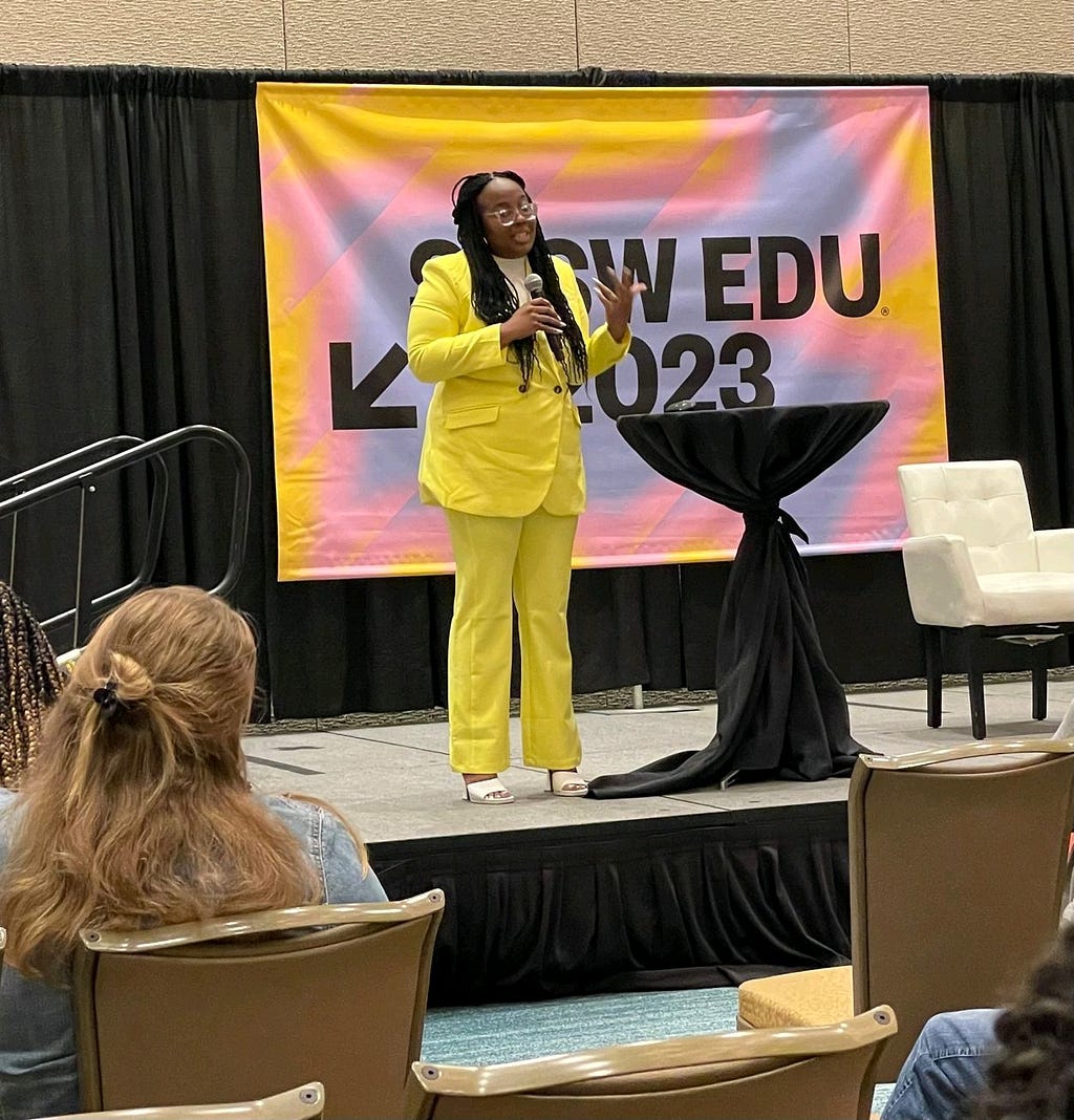 Image of Kara speaking on a stage to a diverse audience at the SXSW EDU meeting. Kara has a yellow suit on with whit open-toed heels. Her hair is in braids with the sides pulled back. There is a yellow, blue, and pink SXSW EDU 2023 banner behind her. On stage, there is a black podium-like table and white chair next to her.