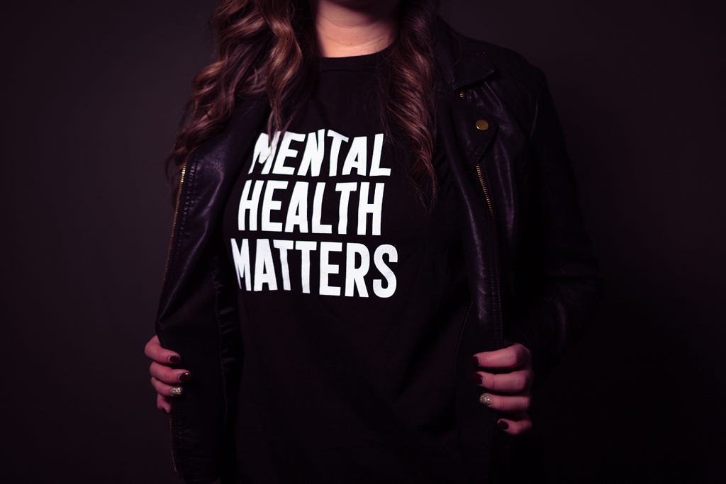 A woman standing against a dark grey background holds her leather jacket open to reveal a black shirt that says ‘Mental Health Matters.’