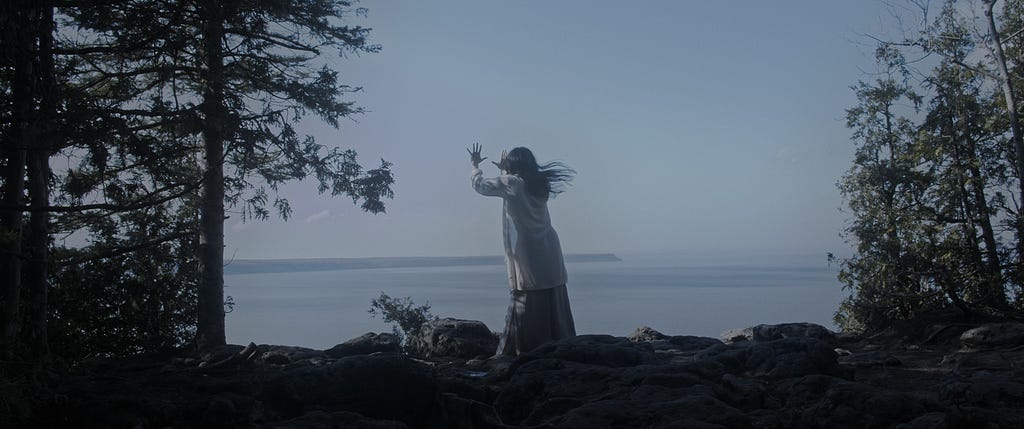 Dancer and co-director Kin Nguien stands at the edge of a forested landscape, facing the water hundreds of feet below. Her hands are raised in front of her with her palms towards the sky in front of her.