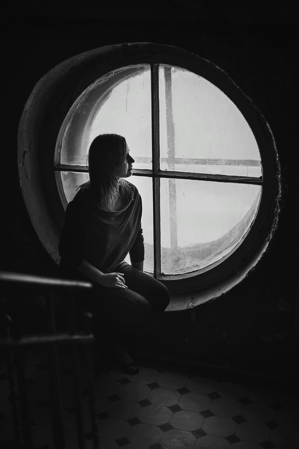 Grayscale photo of a woman sitting near a round wooden framed window