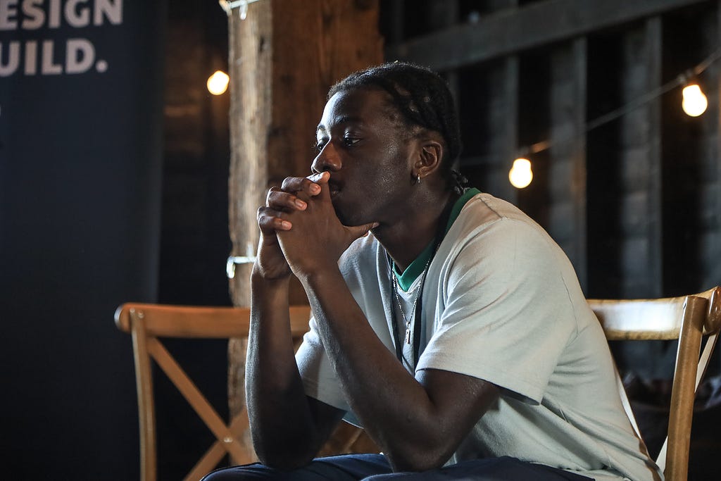 Male guest sits with his elbows on his knees and his chin in his hands. He wears a white t-shirt with a green trim around the neck. He sits on a wooden chair and there is a small glimpse of the Black Design Guild banner and logo behind him.