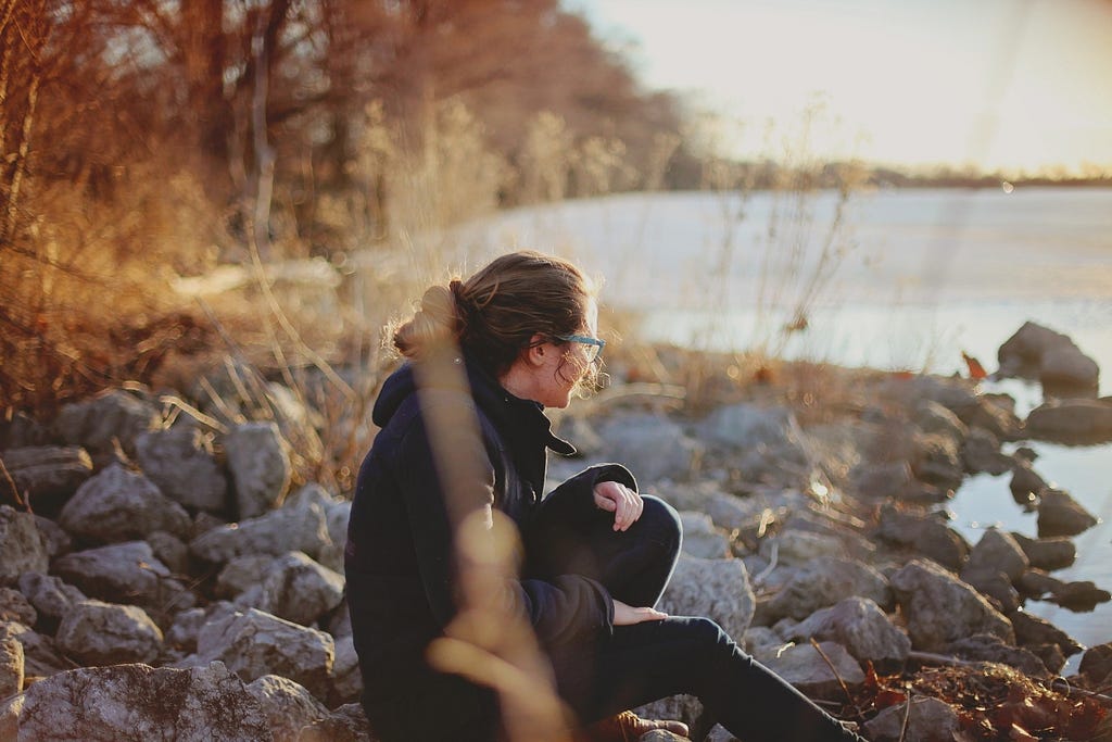A woman sitting on a rocky riverbank alone, looking over the water