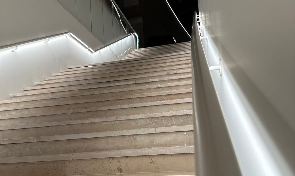 A modern staircase at the Musée de la Marine ascends from the model room to the main floor. The steps are made of textured beige stone, edged with white LED lights enhancing visibility. A sleek white handrail runs alongside, reflecting the light, contributing to a clean, contemporary design