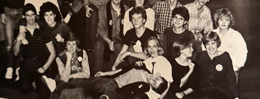According to ChatGPT, this is a black-and-white photo of a group of high school students posing together, likely from the 1980s. They are arranged in several casual rows, smiling and leaning against each other, with one student lying playfully across the front. The scene conveys youthful camaraderie and nostalgia.