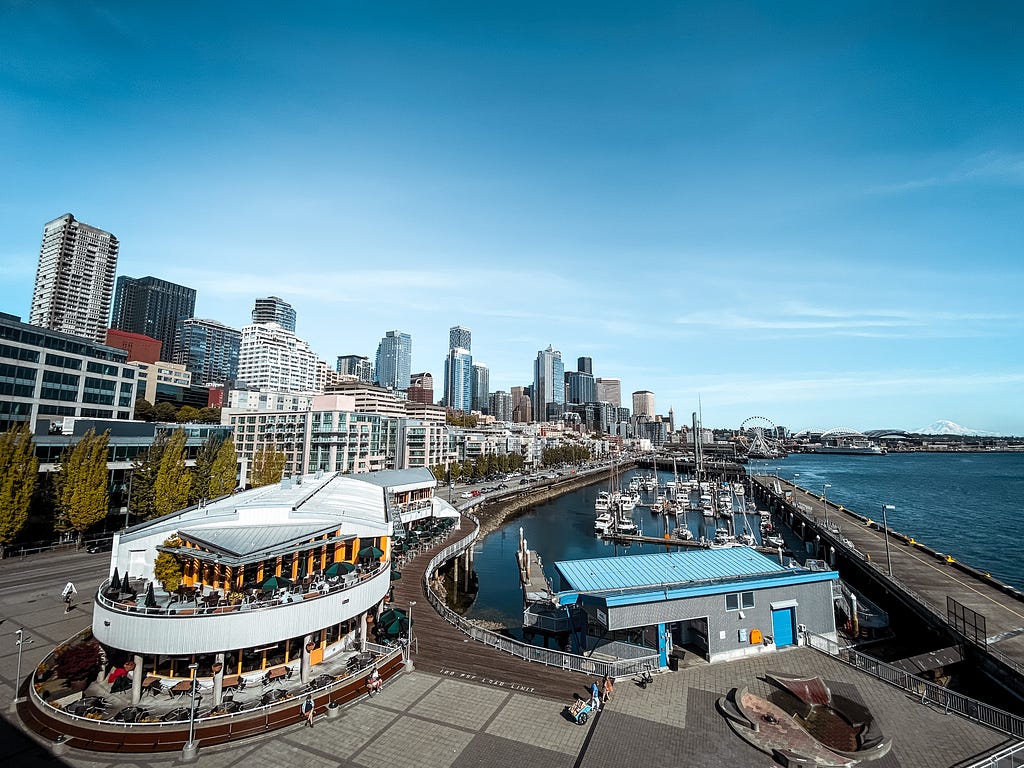 A view of the Seattle waterfront under blue skies, the cityscape to the left and Puget Sound to the right. Tahoma (Mount Rainier) is visible in the distance.