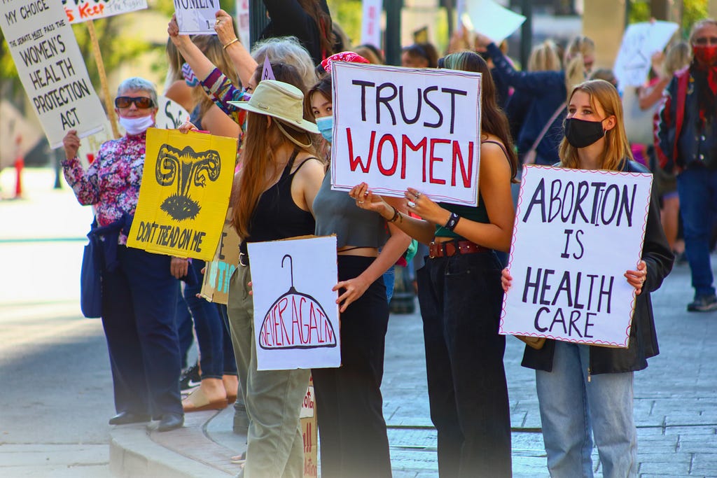 Women on a street holding signs including those that read ‘Trust Women’ and ‘Abortion is Health Care’