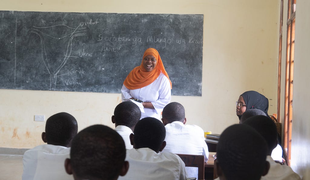 Health worker standing in a classroom in front of a chalkboard teaching students about HPV and the importance of vaccinations.