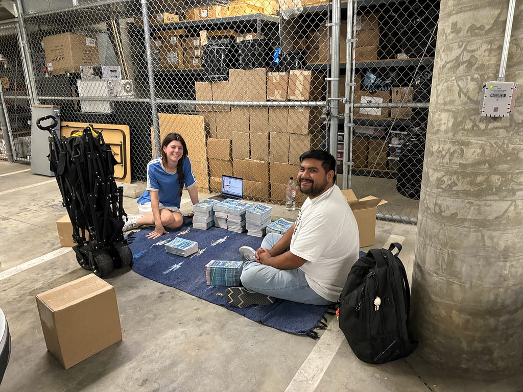 Two people surrounded by boxes, packing and stacking blue booklets.