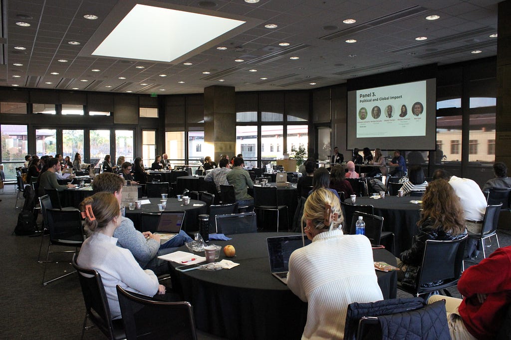 Photo of large conference room with people sitting around a dozen round tables. At the front of the room is a large screen that reads “political and global impact” with five panelists seated under it.