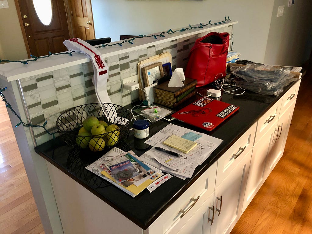 Author’s photo of a kitchen counter with piles of papers, a laptop, red purse, basket of apples