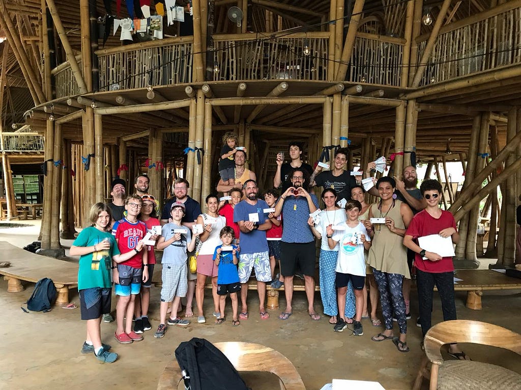 Full body group shot of 20+ students & teachers multi-races pose smiling together inside bamboo structure holding stickers