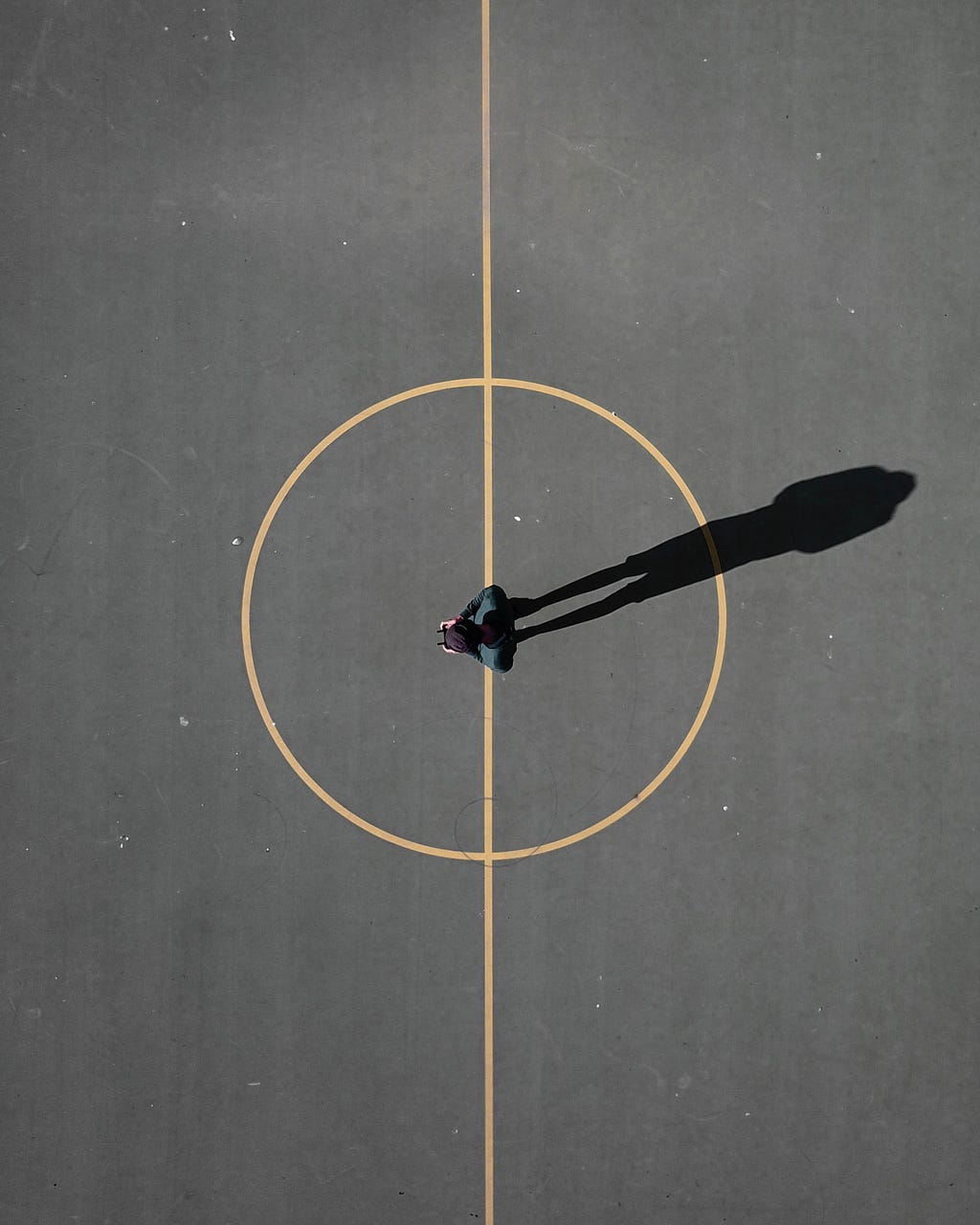 An aerial photo of a person stood in the centre of a tarmac basketball court. A long shadow is cast by the sunlight, which projects the figure’s shadow.