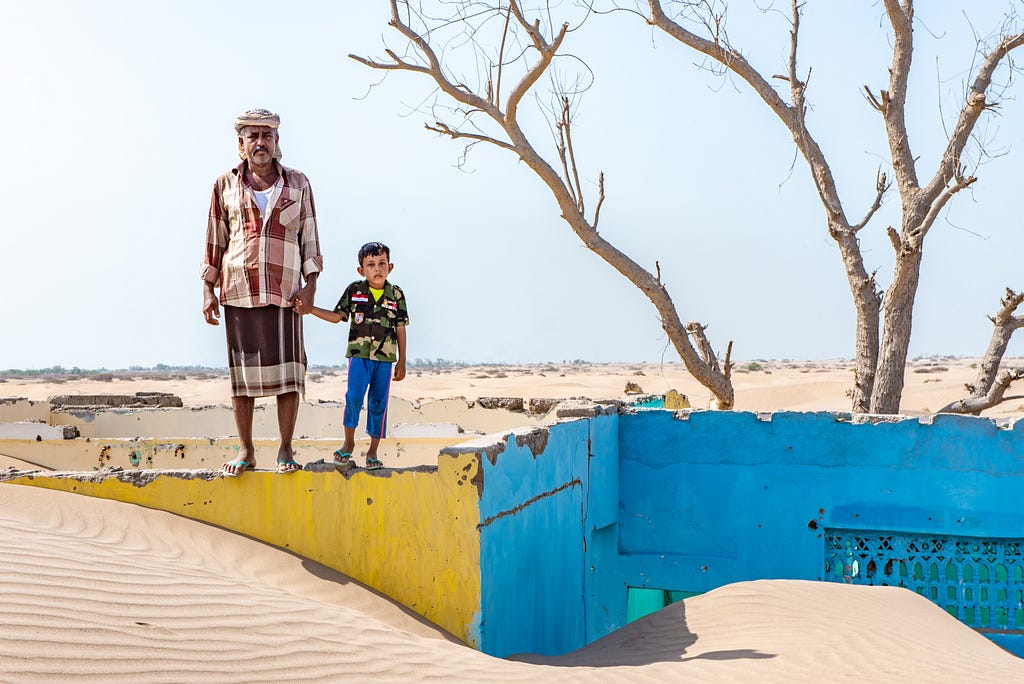 Photo of a father and son holding hands standing on a yellow wall of a house that has lost its roof and has been submerged under sand.