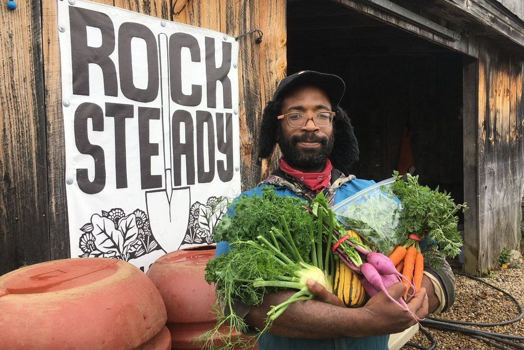 Farm crew holds vegetables in arms standing outside a wooden barn with the Rock Steady sign.