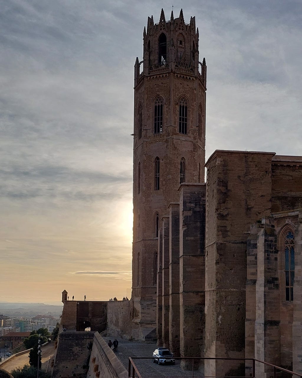 Side face of Lleida’s Seu Vella, with bell tower views. The monument is made of stone, and the sun is shining behind it, providing a beautiful contrast of colors. In the background, you can see the city, which seems very small due to the hill’s height.