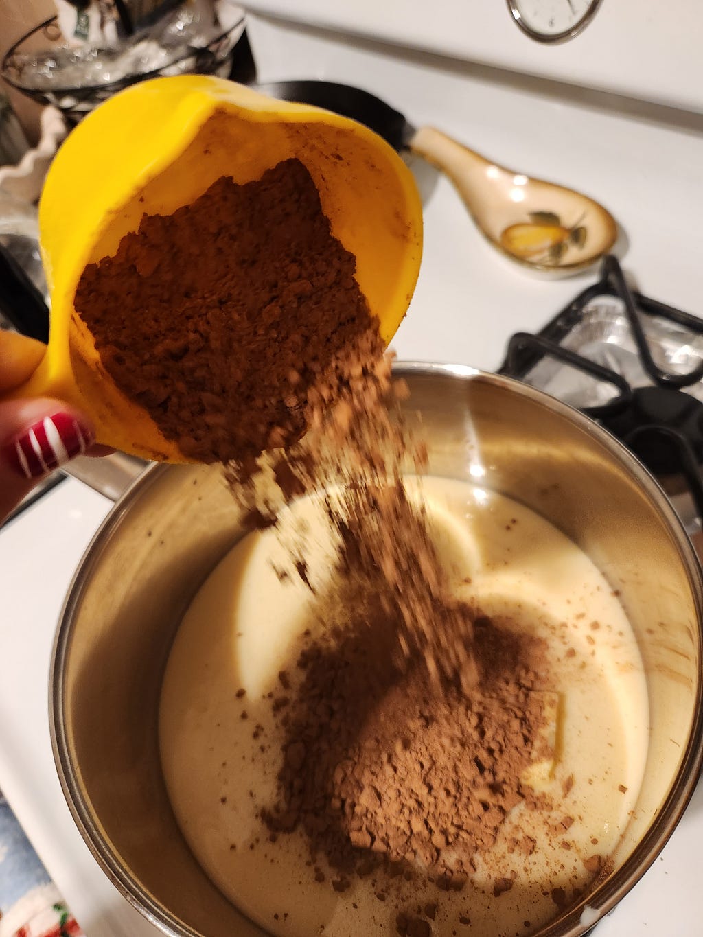 A photo of my hand (thumb visible) pouring in cocoa powder into the condensed milk and butter mixture for brigadeiros. Cocoa powder is falling from a yellow measuring cup into a metal pan on top of a stove.