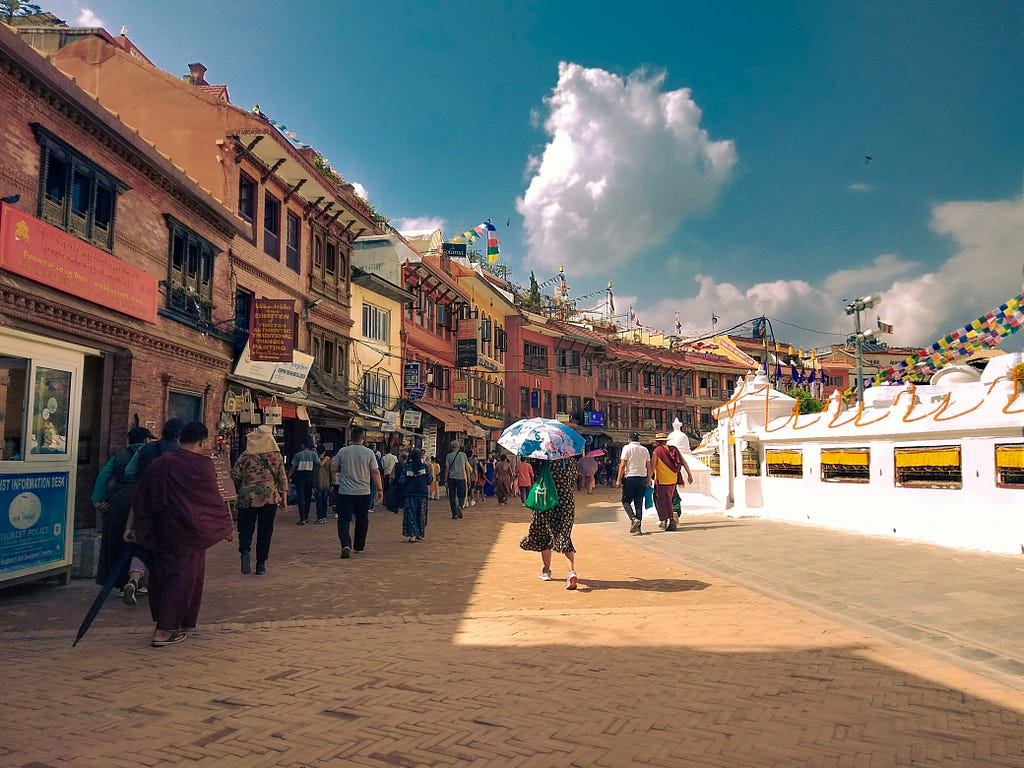 A street in Boudhanath near the Stupa — various shades of brown buildings are on the right side of the photo and the bright white and gold of the stupa on the left with a dusty pedestrian street in the middle. Several people are walking in the middle of the street.