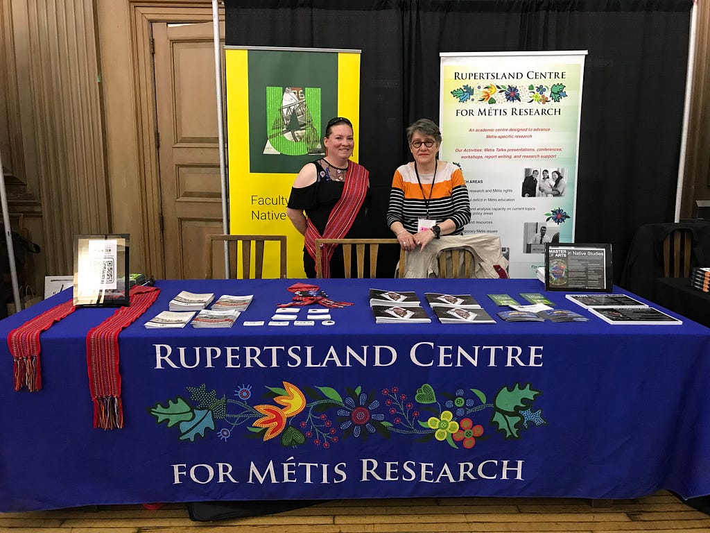 2 women, Amanda Evans and Nathalie Kermoal, stand behind a table that reads “Rupertsland Centre for Metis Research” and in front of banners for the Faculty of Native Studies and RCMR.